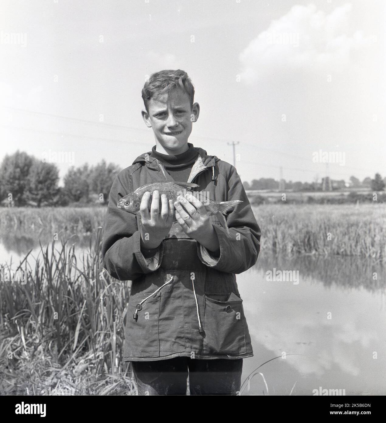 1960s, historisch, ein Teenager-Junge mit Kapuzenoberteil, Seitentaschen und Kordelzug am Bund, der an einem Flussufer steht und seinen Fang zeigt, England, Großbritannien. Die Jugend nahm an einem Fischfang Teil, Stockfoto