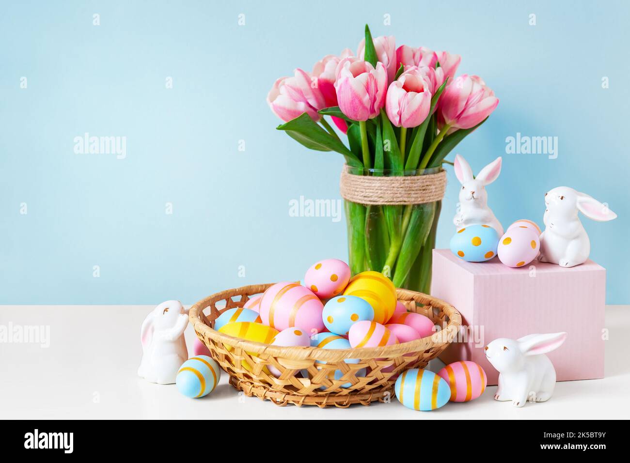 Ostern und Frühlingsferien Grußkartenkonzept. Rosa Tulpen, osterhasen mit bunten Eiern im Korb und blauem Hintergrund. Bemalte Ostereier Stockfoto