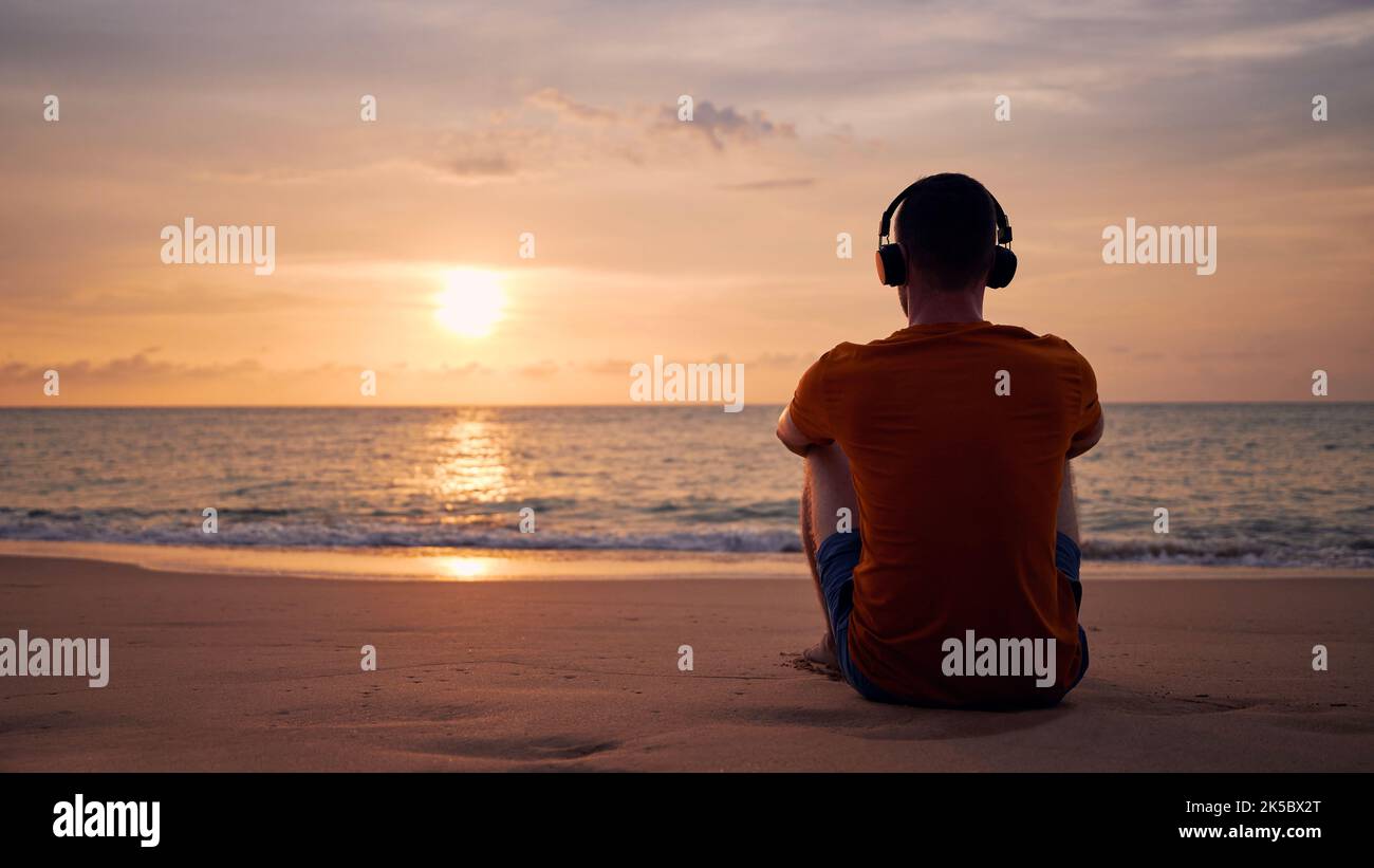Rückansicht des Mannes mit Kopfhörern am Strand. Gelassenheit, Kontemplation und Musikhören bei schönem Sonnenuntergang. Stockfoto
