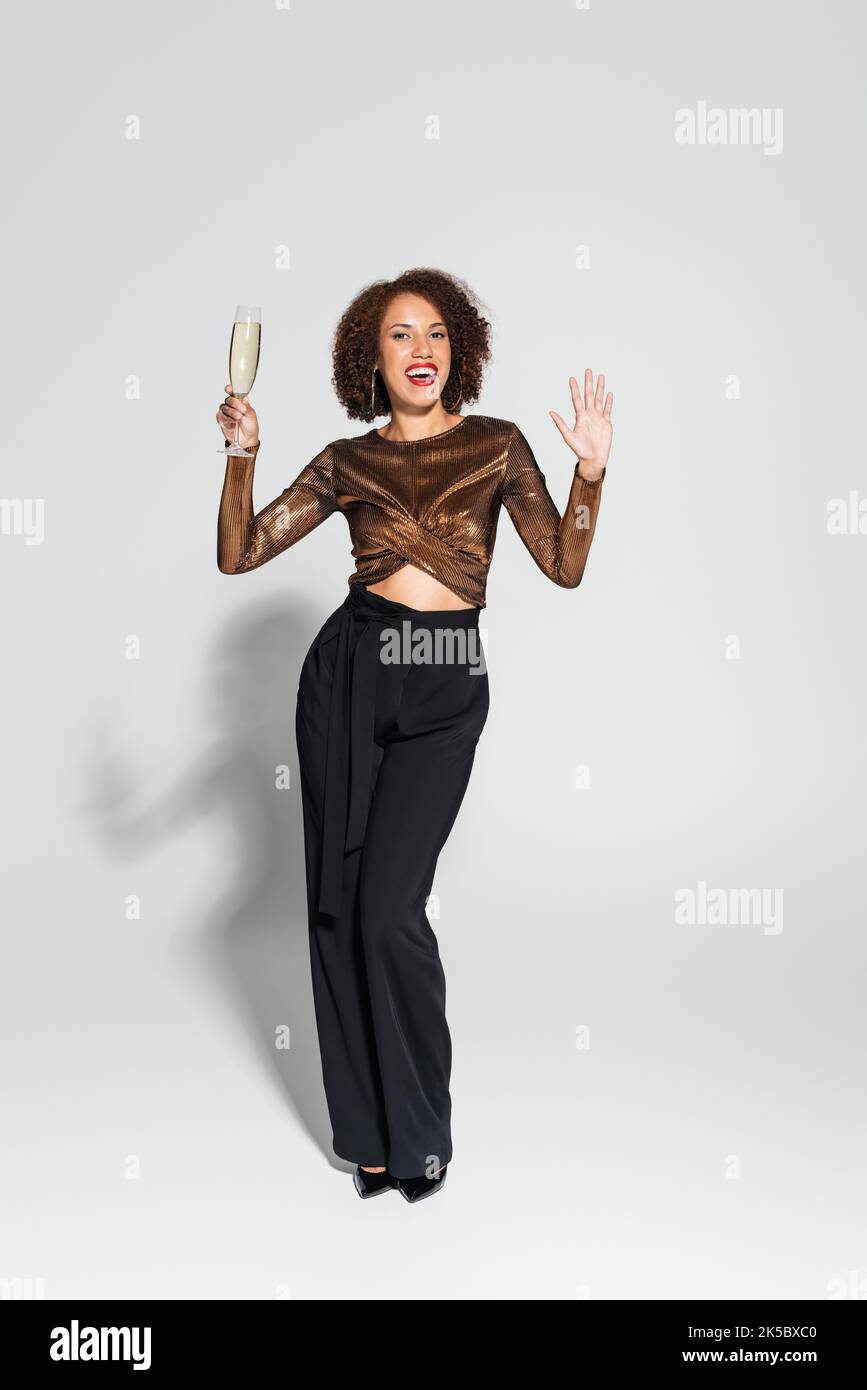 Volle Länge der fröhlichen und stilvollen afroamerikanischen Frau mit Champagner und winkende Hand auf grauem Hintergrund, Stockbild Stockfoto