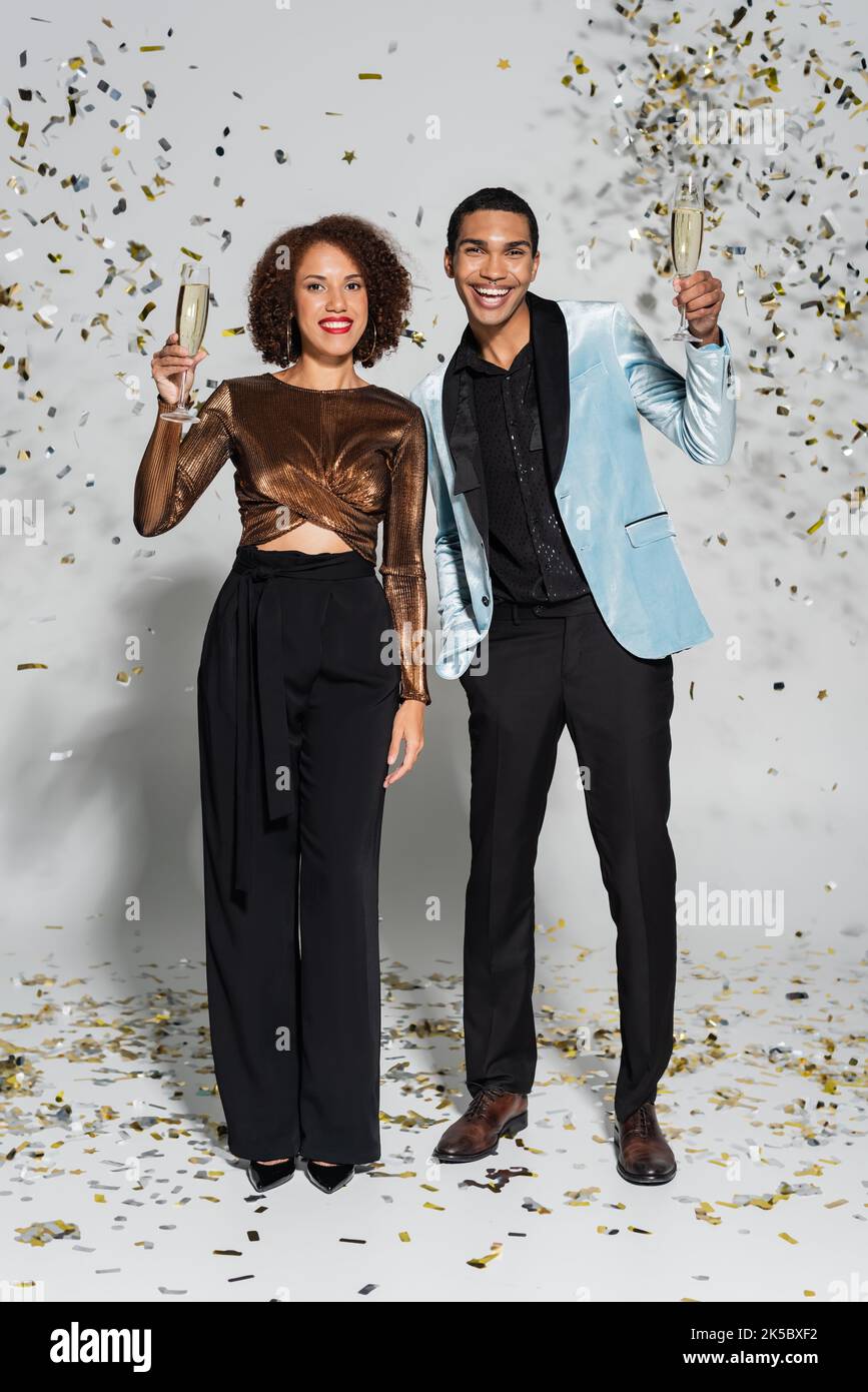 Glückliches afroamerikanisches Paar in festlichen Kleidern mit Champagner in der Nähe von Konfetti auf grauem, stockem Bild Stockfoto