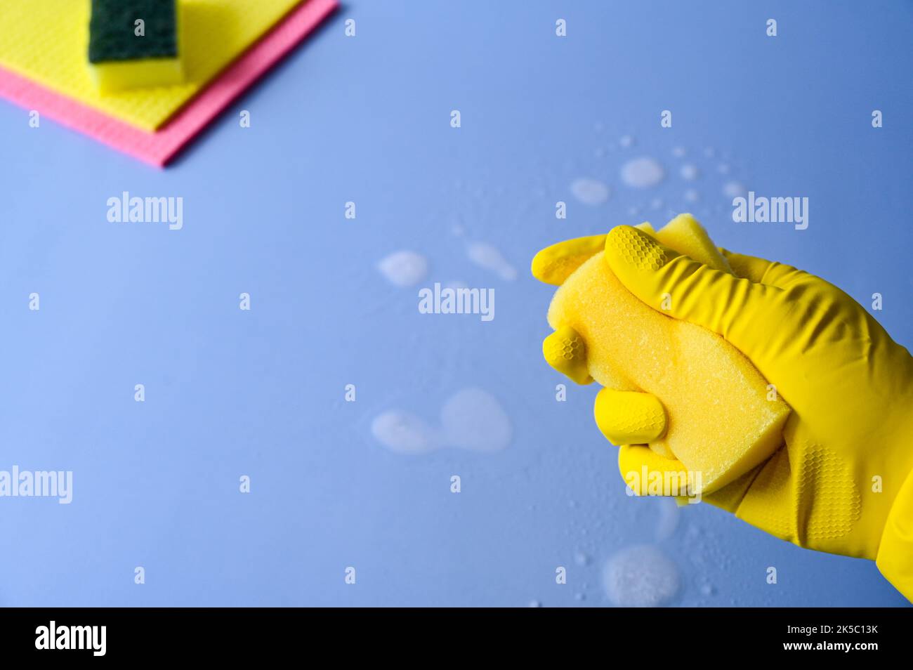 Hand mit gelbem Gummihandschuh hält einen Reinigungsschwamm auf blauem Hintergrund. Stockfoto