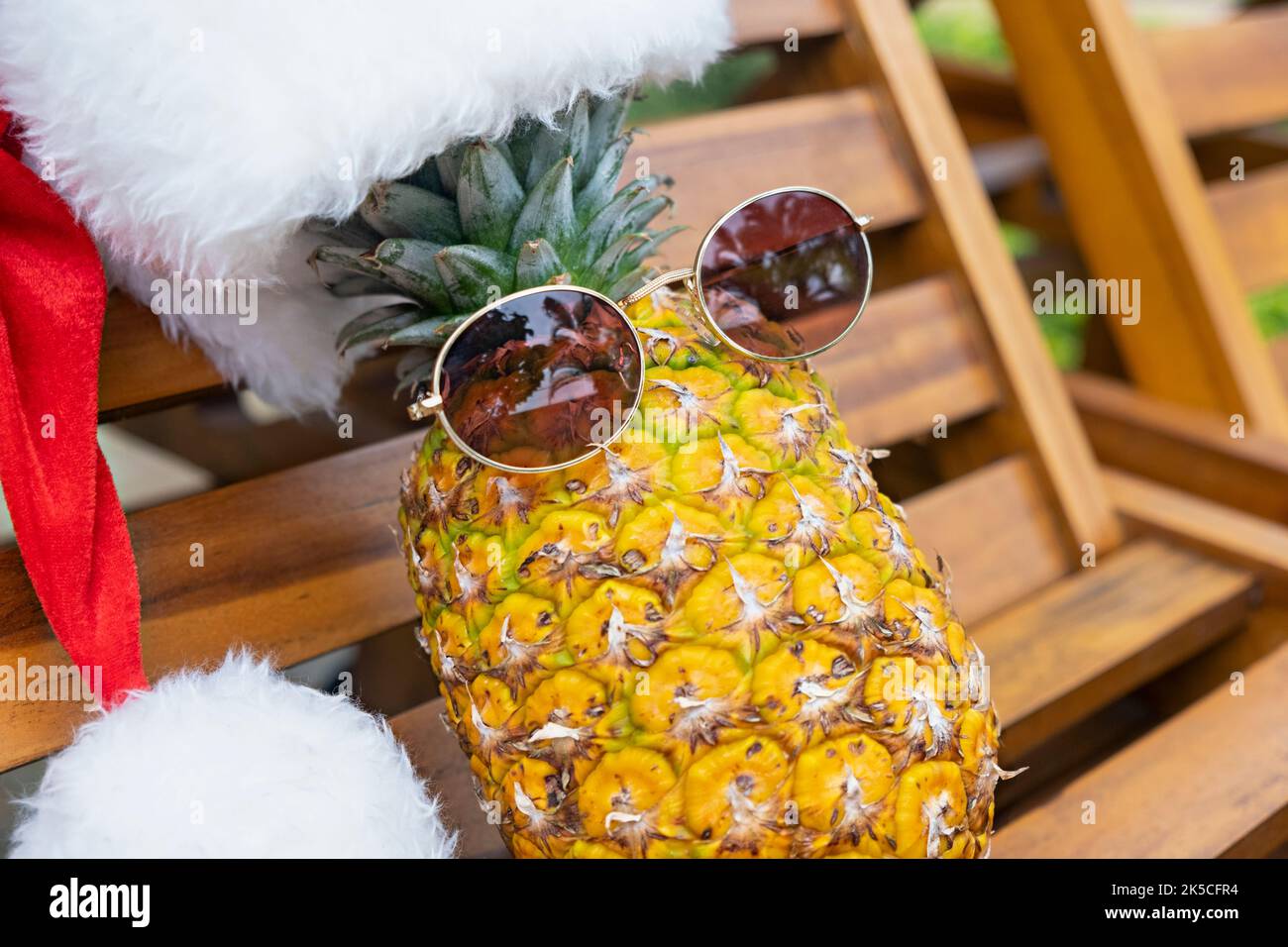Nahaufnahme einer entspannenden, lustigen Ananas mit Sonnenbrille und Weihnachtsmann-Hut liegt bei sonnigem Wetter in den Tropen auf einer hölzernen Sonnenliege. Weihnachten in Stockfoto