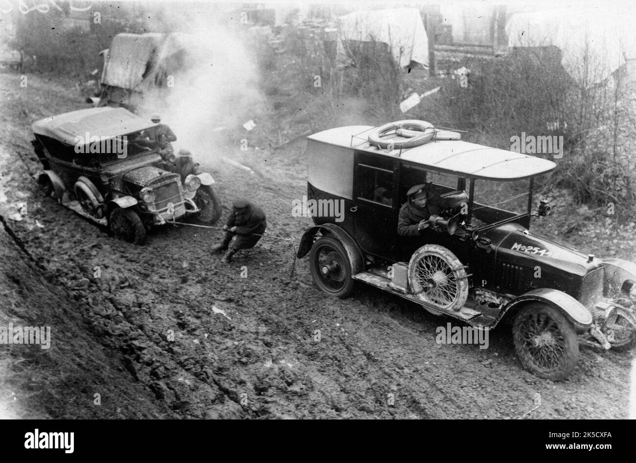 Auto wird geschleppt, Frankreich, während des Ersten Weltkriegs.Dieses Bild zeigt ein Auto, das durch dicken Schlamm geschleppt wird. Als der Wagen vorne versucht, den anderen freizuziehen, gibt ein Soldat eine Hand, indem er am Schleppseil zieht. Eine Gruppe von Soldaten, die neben dem unbeweglichen Auto stehen, tragen zum Kampf bei. Es scheint ein Lager direkt hinter der Straße zu geben, auf der sie sich befinden. Das Fahrzeug vorne ist mit der Nummer „M25446“ auf der Motorhaube bemalt. Extreme Wetterbedingungen, Überschwemmungen und Schlamm, die sich daraus ergaben, verursachten enorme Probleme und Gefahren für die Soldaten an der Front. Stockfoto