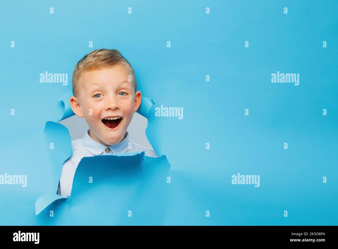 Happy cute boy hat Spaß auf blauem Hintergrund Wand gespielt, klettert durch ein Loch in das Papier. Helle und lustige Emotionen des Jungen Stockfoto