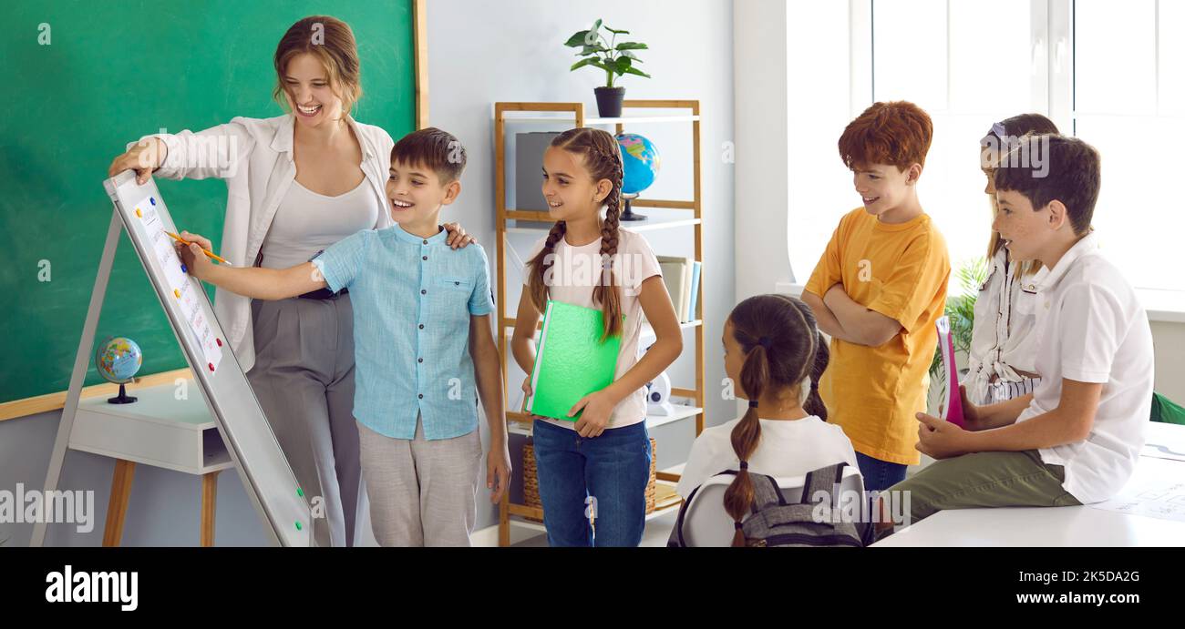 Fröhliche Kinder erfüllen die Aufgabe in der Nähe des Flipchart zusammen mit der Lehrerin in der Grundschule Stockfoto