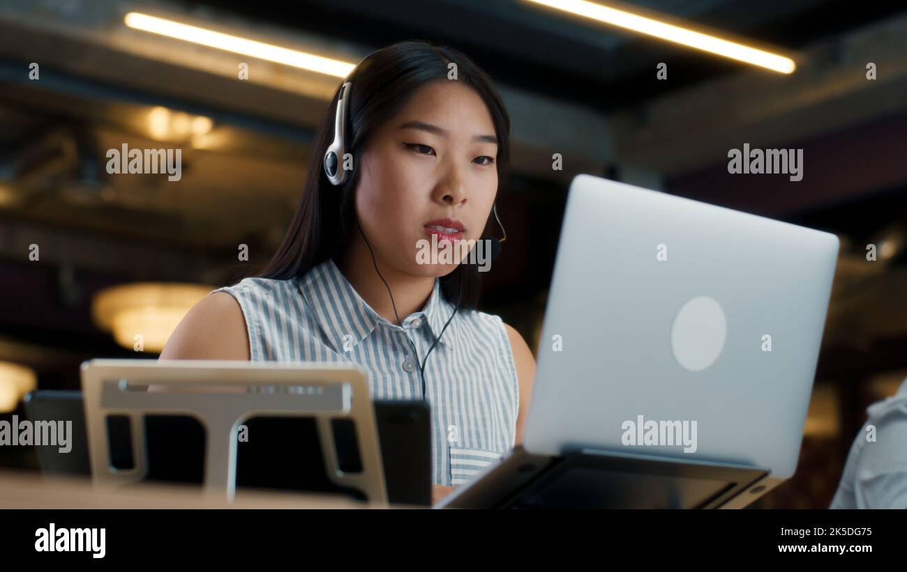 Asiatische Sprecherin im Headset, die online mit dem Kunden spricht, mit einem Laptop und einem digitalen Tablet. Arbeiten im Callcenter des Marketingunternehmens Stockfoto