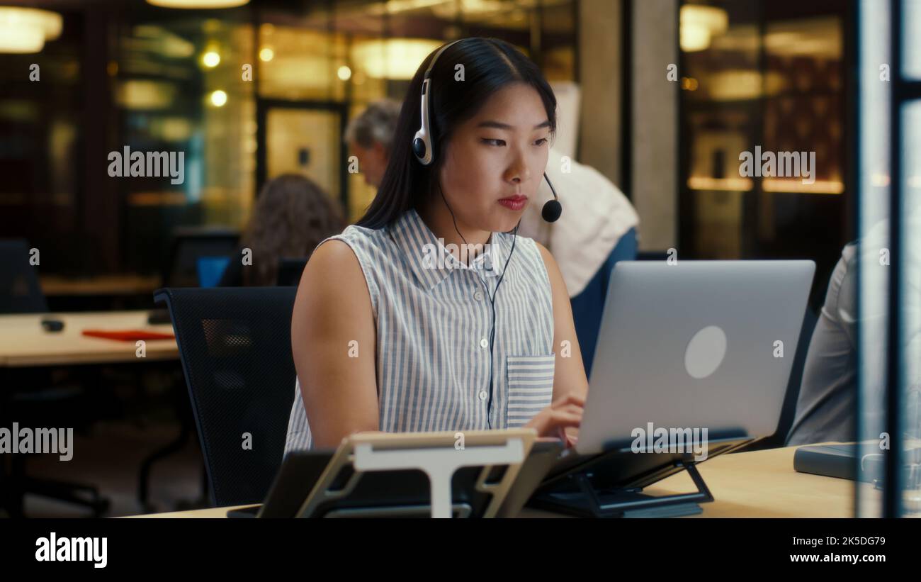 Asiatische Frau im Headset, die mit dem Kunden spricht, mit einem Laptop und einem digitalen Tablet. Weibliche Betreiberin, die im Callcenter-Büro des Marketingunternehmens arbeitet Stockfoto