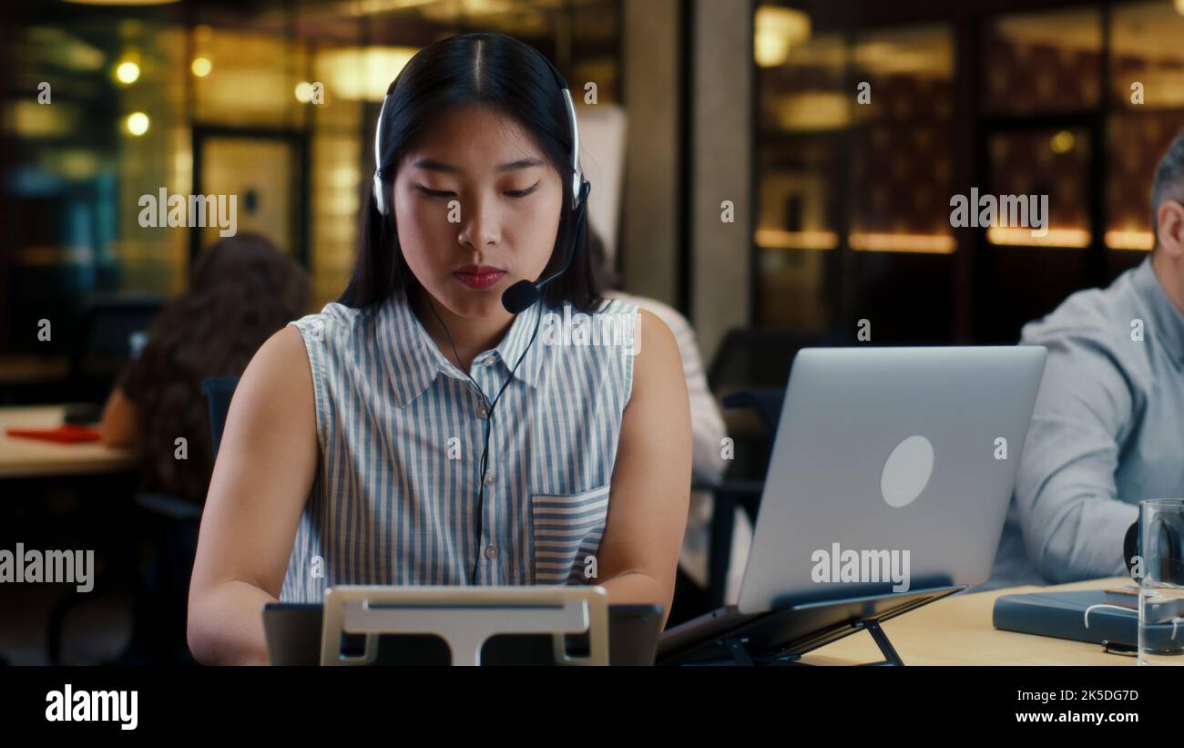 Asiatische Frau im Headset, die mit dem Kunden spricht, mit einem Laptop und einem digitalen Tablet. Weibliche Betreiberin, die im Callcenter-Büro des Marketingunternehmens arbeitet Stockfoto