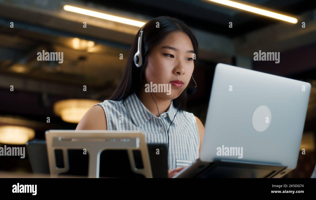 Asiatische Sprecherin im Headset, die online mit dem Kunden spricht, mit einem Laptop und einem digitalen Tablet. Arbeiten im Callcenter des Marketingunternehmens Stockfoto