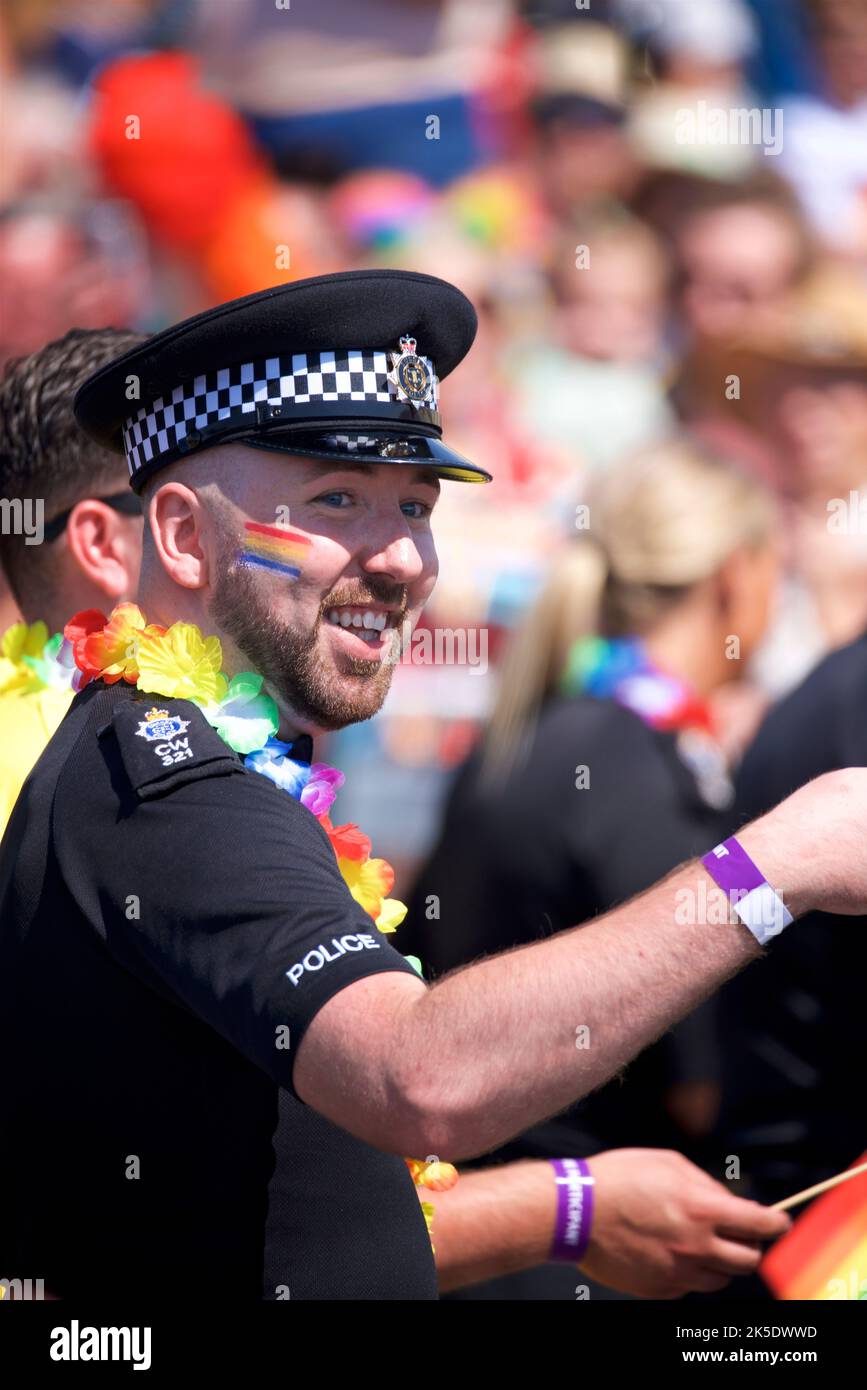 Brighton & Hove Pride Festival, Brighton & Hove, East Sussex, England. Britischer Polizist in Uniform mit Regenbogenfahne auf dem Gesicht gemalt und mit der Hand gewellt Stockfoto