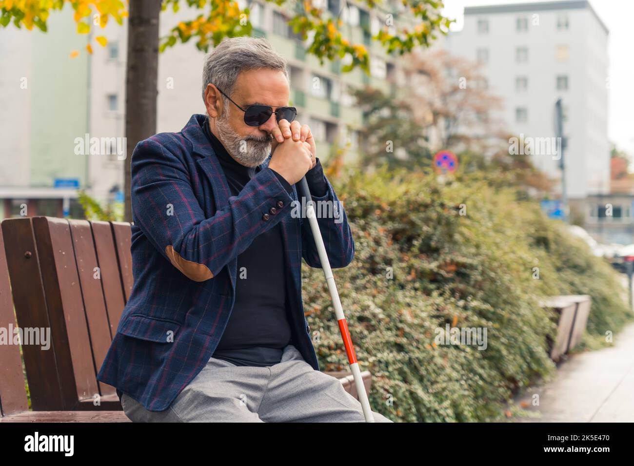 Kaukasischer grauhaariger, erwachsener, gut aussehender Mann in Sonnenbrille mit vollständigem Sichtverlust, der auf einer Bank im Park sitzt und weißen Stock neben seinem Gesicht hält. Außenaufnahme. Hochwertige Fotos Stockfoto