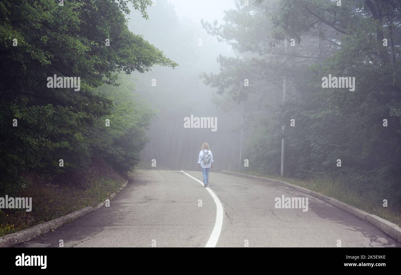 Straße in nebligen Wald, einsame Mädchen Wanderer geht weg in unheimlichen Wäldern, verloren junge Frau Reisende ist allein im Nebel. Landschaft von gruseligen Ort. Konzept von h Stockfoto