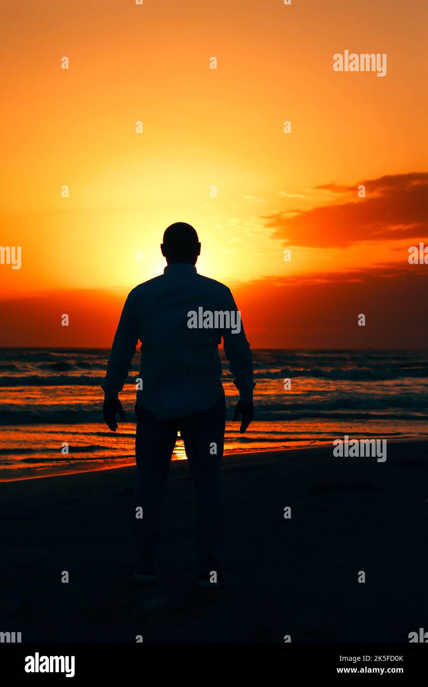 Eine Silhouette eines Mannes, der während des Sonnenuntergangs auf das Meer am Strand blickt und sich auf Entspannung konzentriert. Stockfoto