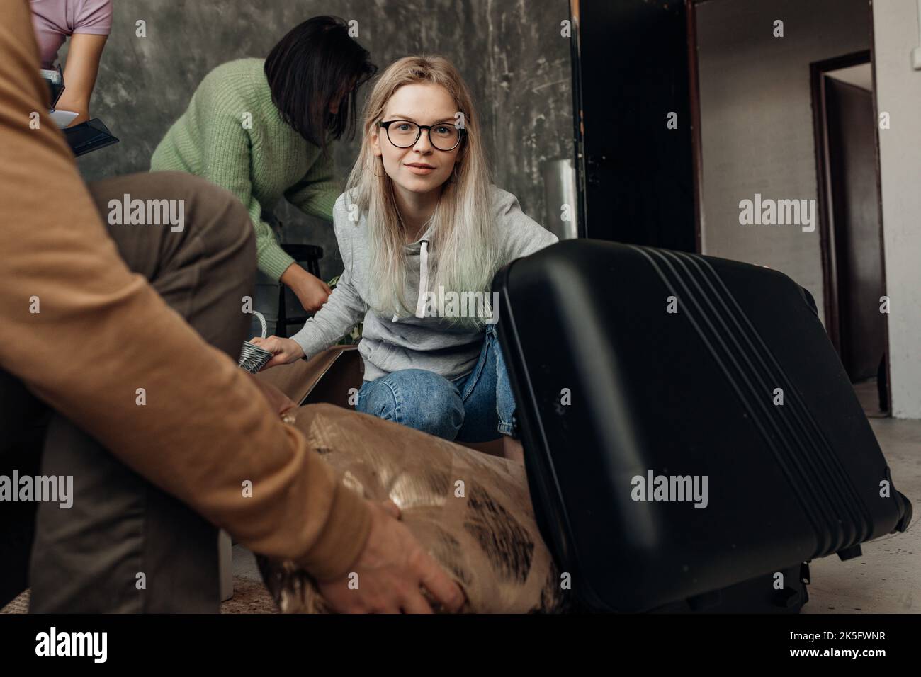 Beschäftigte Frau in Brille mit Freunden packen Koffer und versuchen, Dinge im Wohnzimmer zu passen. Transport Versand Stockfoto