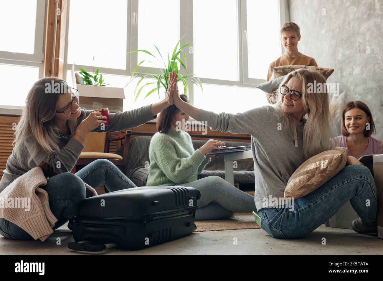 Lächelnde, energische Schülerin, die fünf auf dem Boden gibt, während sie das Gepäck mit Sachen im Wohnzimmer packt und eine Freundin zeigt Stockfoto