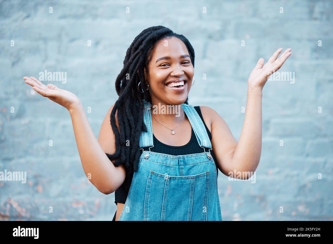 Schwarze Frau, Lächeln und Hände für verwirrte oder unsichere Geste, die draußen vor städtischem Hintergrund auf der Straße gegen die blaue Wand posiert. Lachende Frau Stockfoto