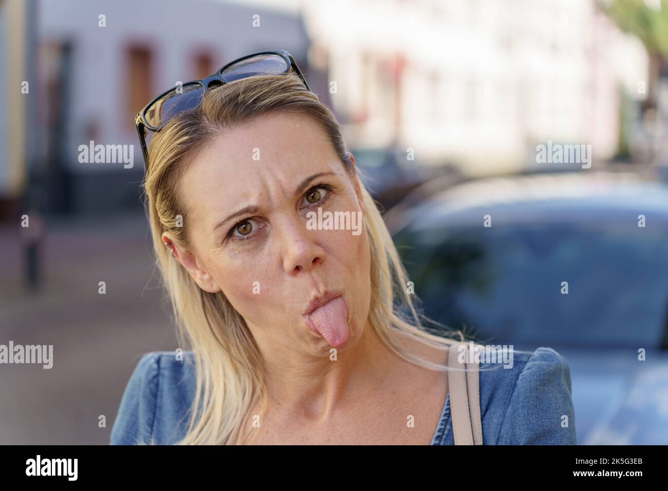 Trendige blonde Frau zieht ein lustiges Gesicht aus ihrer Zunge an der Kamera, während sie zur Kamera schaut Stockfoto