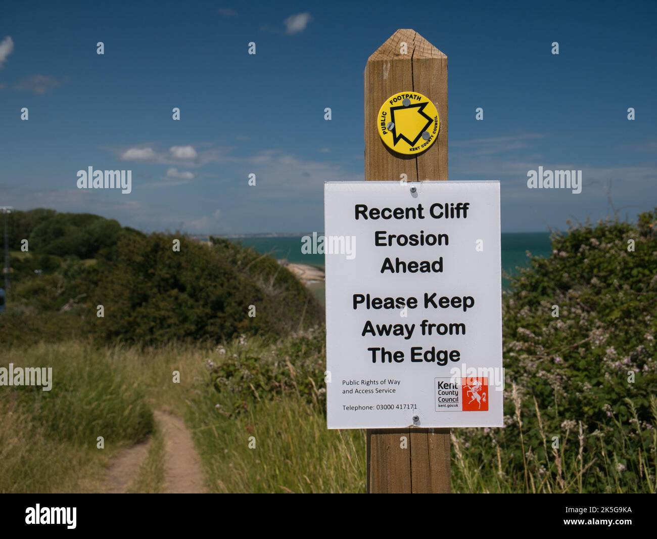 Ein weißes rechteckiges Schild, das an einem Holzpfosten neben einem Pfad befestigt ist, warnt Wanderer vor der jüngsten Erosion der Klippen am Küstenpfad. Aufgenommen an einem sonnigen Tag im Sommer Stockfoto