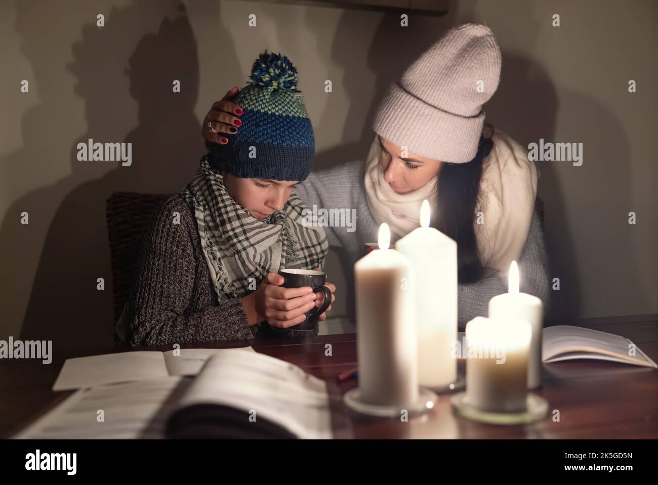 Besorgte Mutter tröstet ihren eiskalten Sohn mit Tee und studiert bei Kerzenlicht in der Kriegszone, beide tragen Winterkleidung zu Hause. Stockfoto