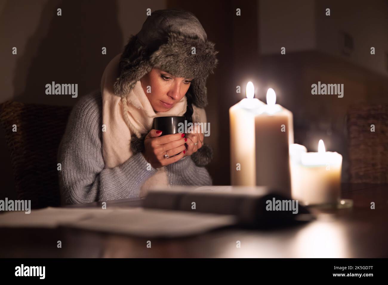 Eine eiskalte junge Frau in Winterkleidung wärmt ihre Hände auf einer Tasse Tee und leuchtet mit Kerzen, während Stromausfälle durch Stromausfälle verursacht werden. Stockfoto