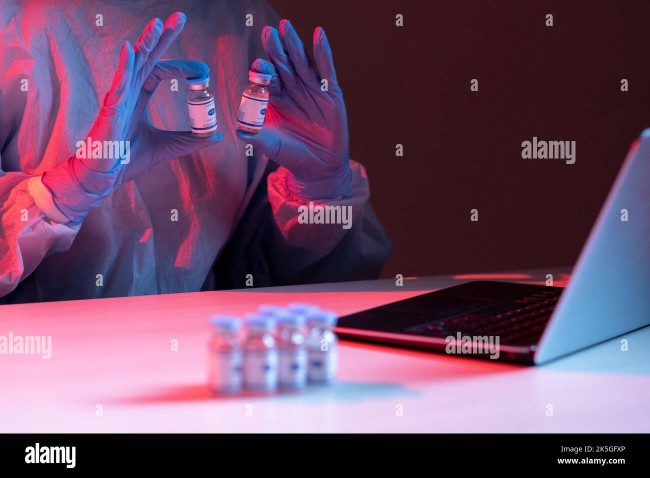 Covid-19-Impfstoff. Online-Recherche. Pandemie-Telemedizin. Wissenschaftlerin in PSA-Handschuhen, die Fläschchendosierungsflaschen auf dem Laptop während der Videoaufnahme in rot b zeigen Stockfoto