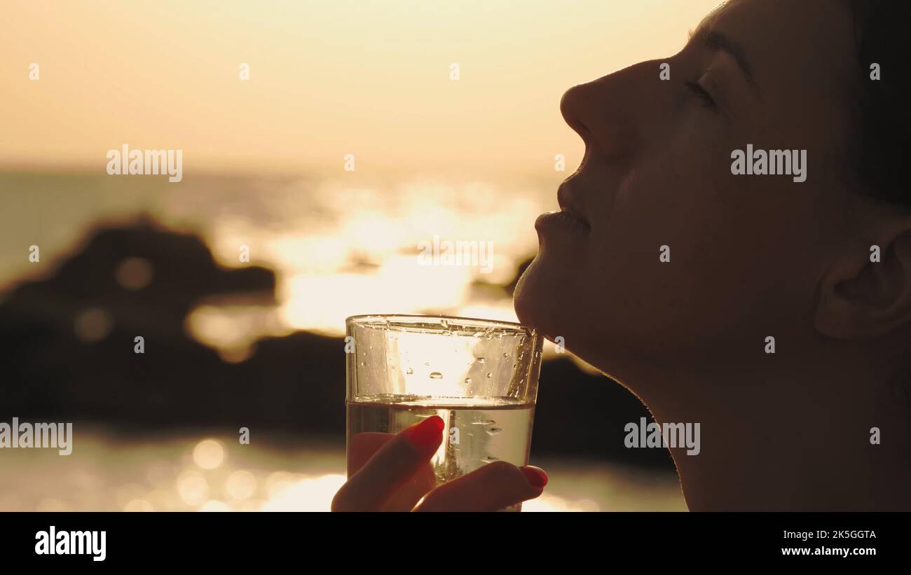 Glas Wasser bei Sonnenuntergang. Nahaufnahme. Silhouette. Frau hält ein Glas klares Wasser in der Nähe ihres Gesichts im Profil. Hintergrund eines Sonnenuntergangs oder Sonnenaufgangs über dem Meer. Hochwertige Fotos Stockfoto