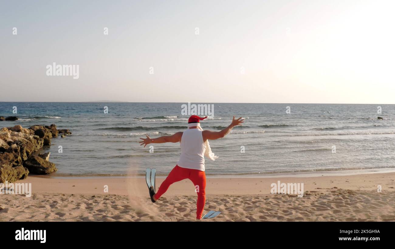 Weihnachtsmann Sommerurlaub. Vater Weihnachten Spaß haben. Lustiger Weihnachtsmann, in Flossen läuft durch Sandstrand zum Meer. Er hat es eilig zu schwimmen, zu schnorcheln. Rückansicht. Hochwertige Fotos Stockfoto