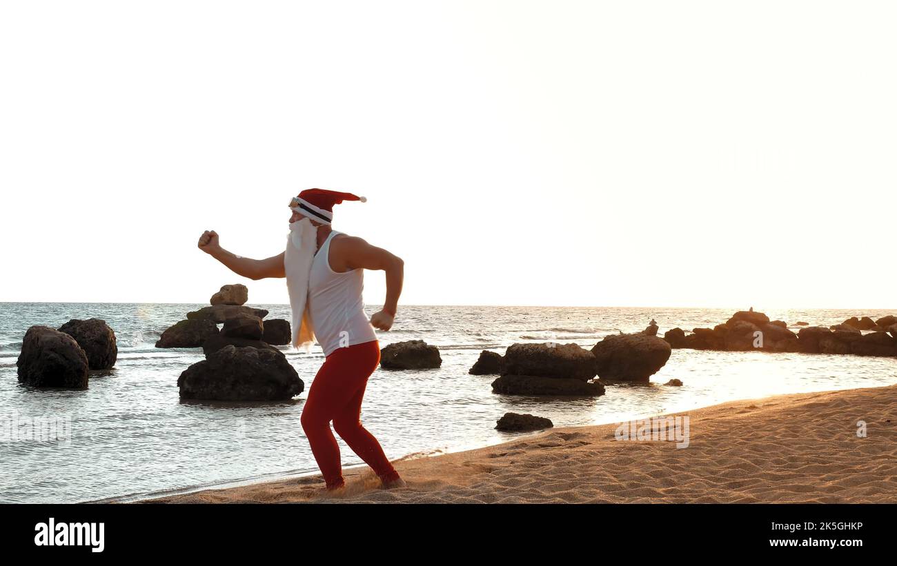 Lustiger weihnachtsmann. weihnachtsmann macht Morgenübungen. weihnachtsmann tanzt am Strand, am Meer, bei Sonnenaufgang. weihnachtsmann ist in T-Shirt und roten Leggings, im Sommerurlaub am Meer. Hochwertige Fotos Stockfoto