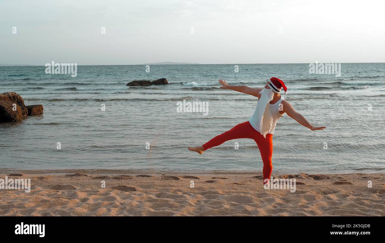 Lustiger weihnachtsmann. weihnachtsmann am Meer, im Sommerurlaub. weihnachtsmann tanzt, springt am Strand, bei Sonnenaufgang. weihnachtsmann in einem T-Shirt und roten Leggings am Meer. Hochwertige Fotos Stockfoto