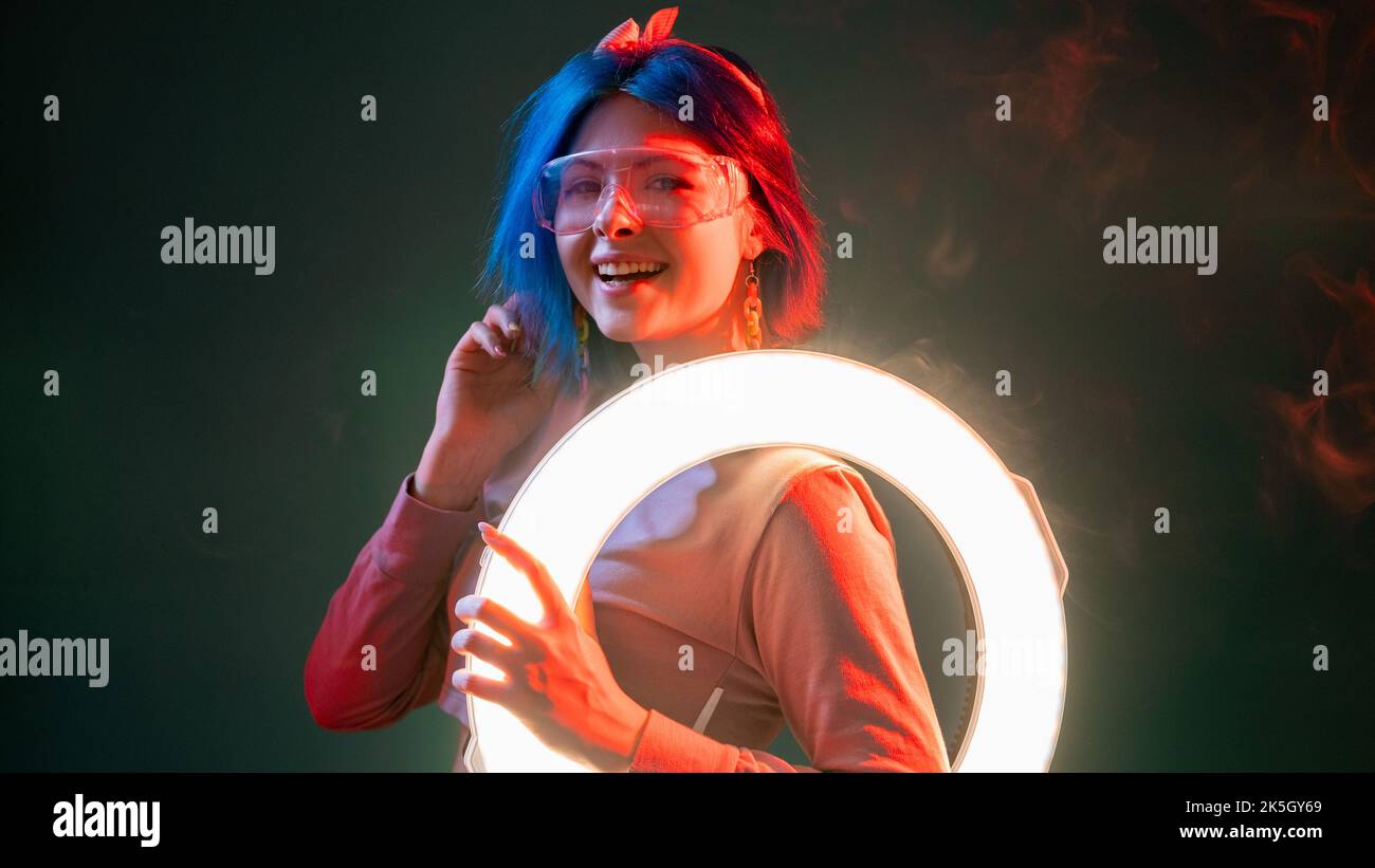 Menschen mit Neonlicht. Futuristischer Style. Moderne Beleuchtung. Fröhlich lächelnde Cyberpunk-Frau mit LED-Ringblitzlampe in rotem Glanz isoliert auf dunklem Hintergrund Stockfoto