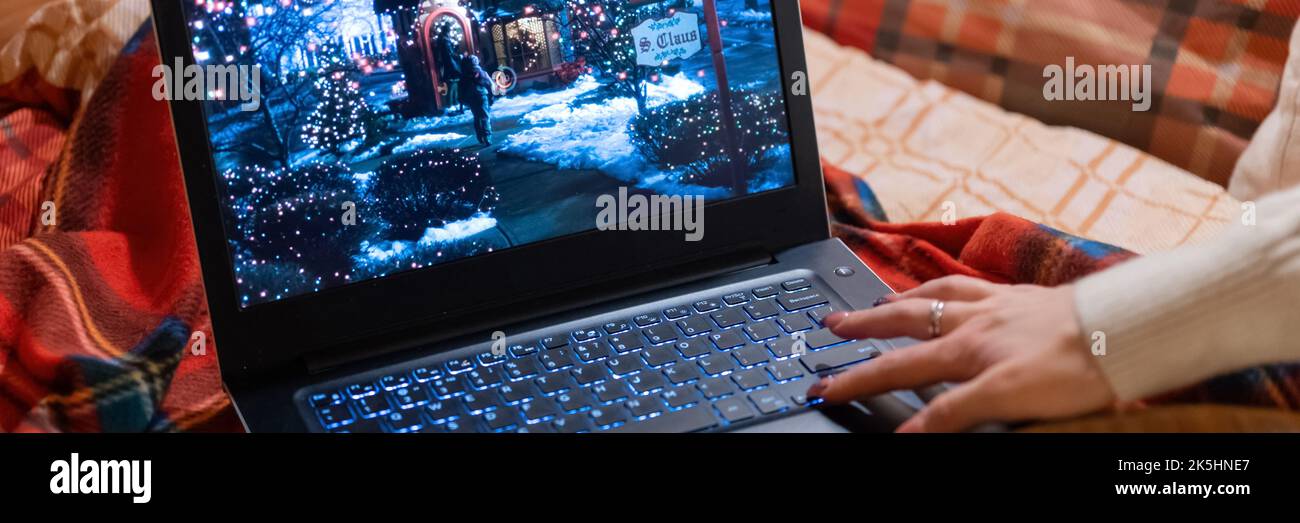 Laptop-Display mit festlichem Weihnachtsfilm „Home Alone“ auf einem roten karierten karierten karierten karierten Bett zu Hause und Video zum weiblichen Handaufzug. Spending Time comi Stockfoto