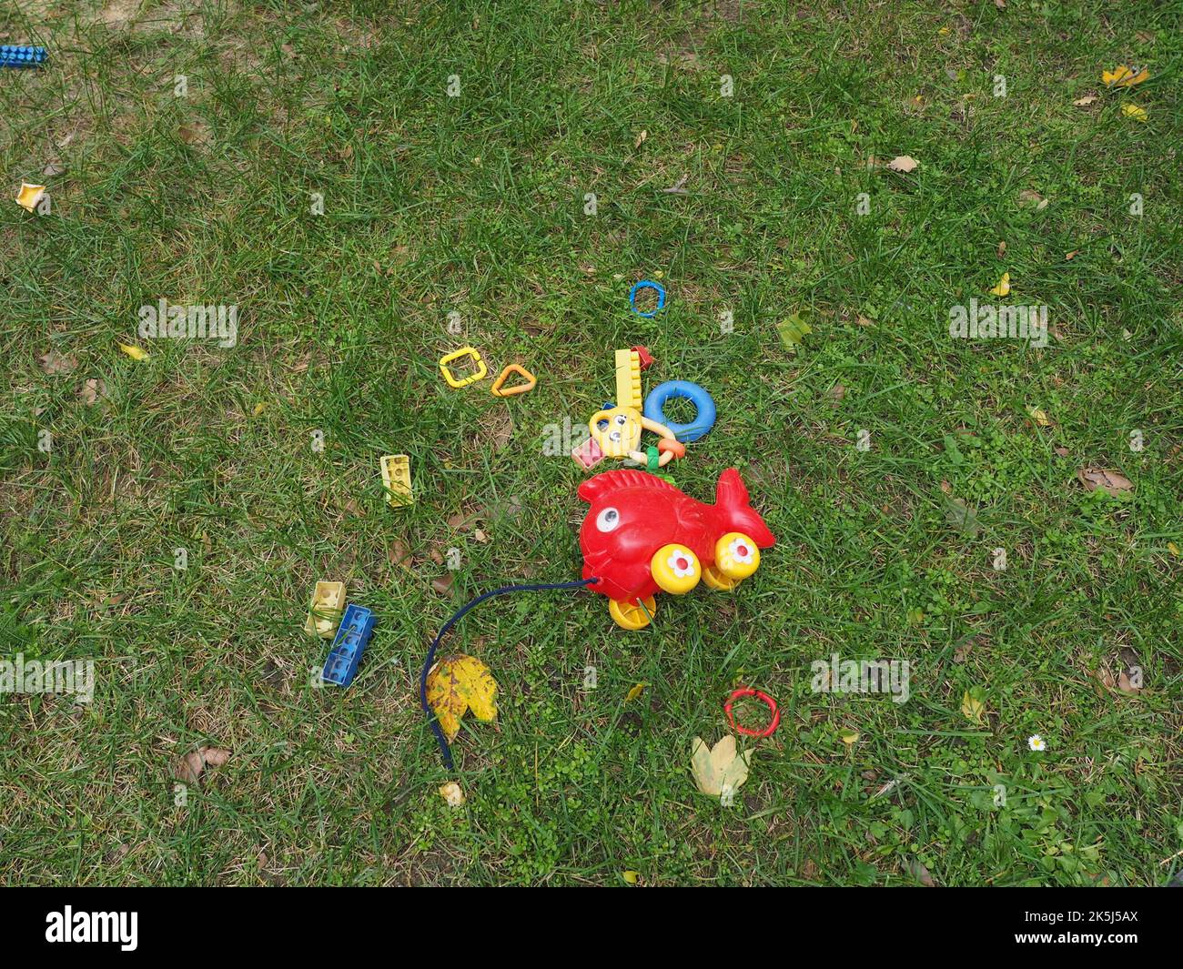 Retro, altes Plastikfischspielzeug auf Rädern mit anderen Plastikspielzeugen, die auf einem Spielplatz im Gras liegen. Spielzeug für Kleinkinder, die von Kindern hinterlassen wurden. Stockfoto