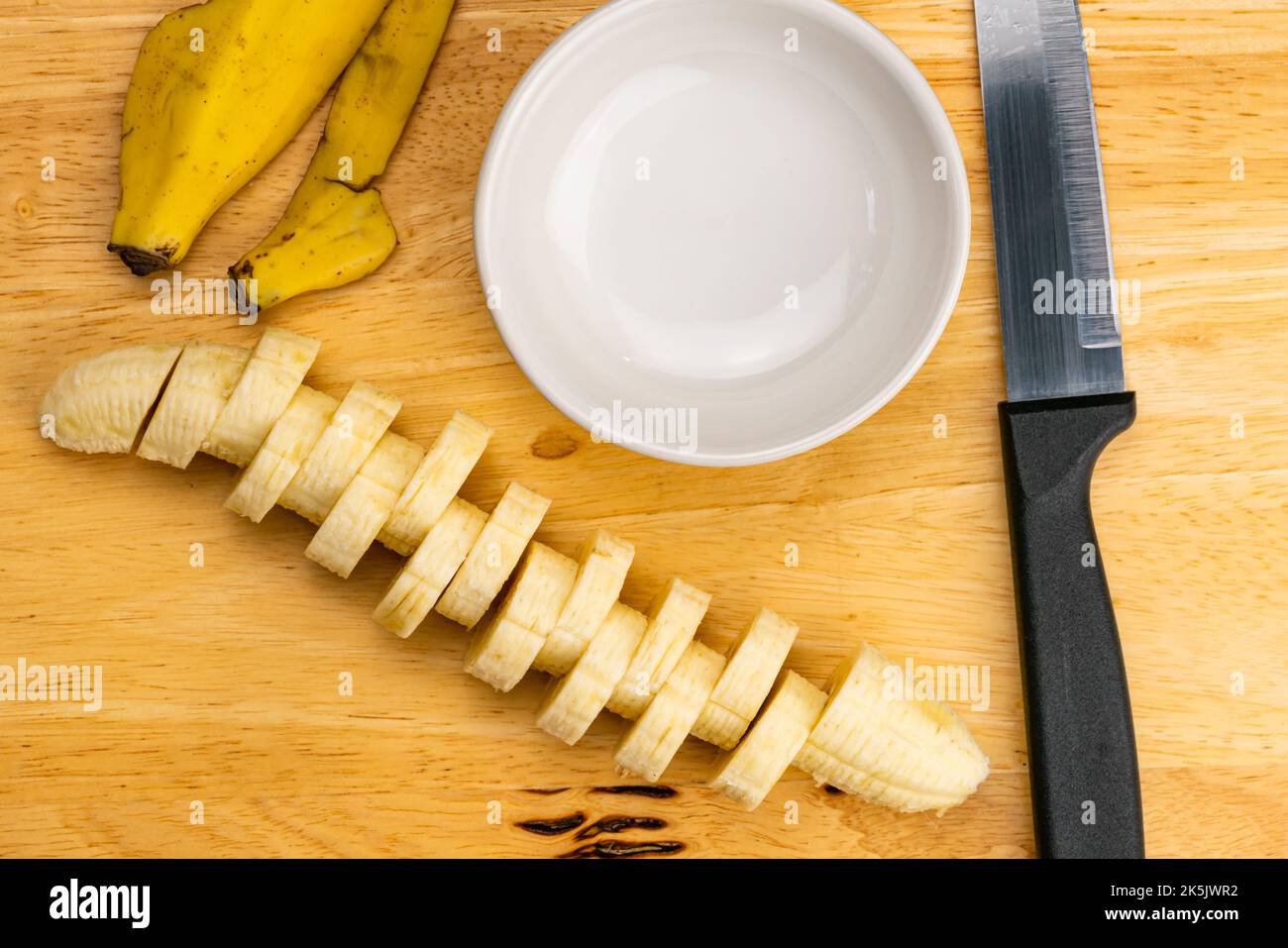 Draufsicht, flache Scheibe roher reifer, organisch geschälter Banane mit Küchenmesser und weißem Keramikbecher auf Holzschneidebrett. Stockfoto