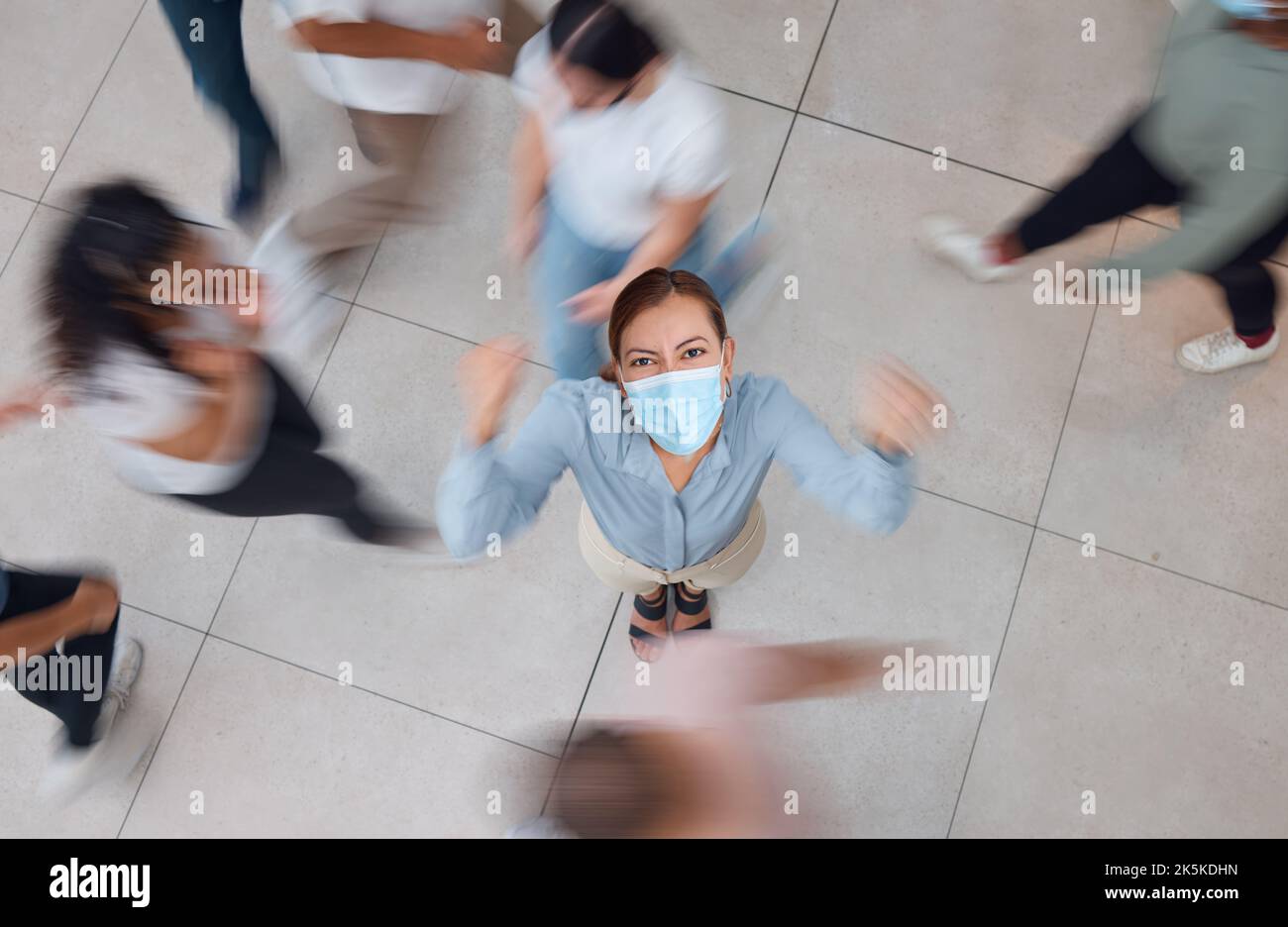 Verärgerte Geschäftsfrau mit einer Maske für Covid und Menschen, die um sie herum laufen. Junges Mädchen mit Gesichtsmaske in einer Menge, frustriert und schüttelt ihre Faust Stockfoto