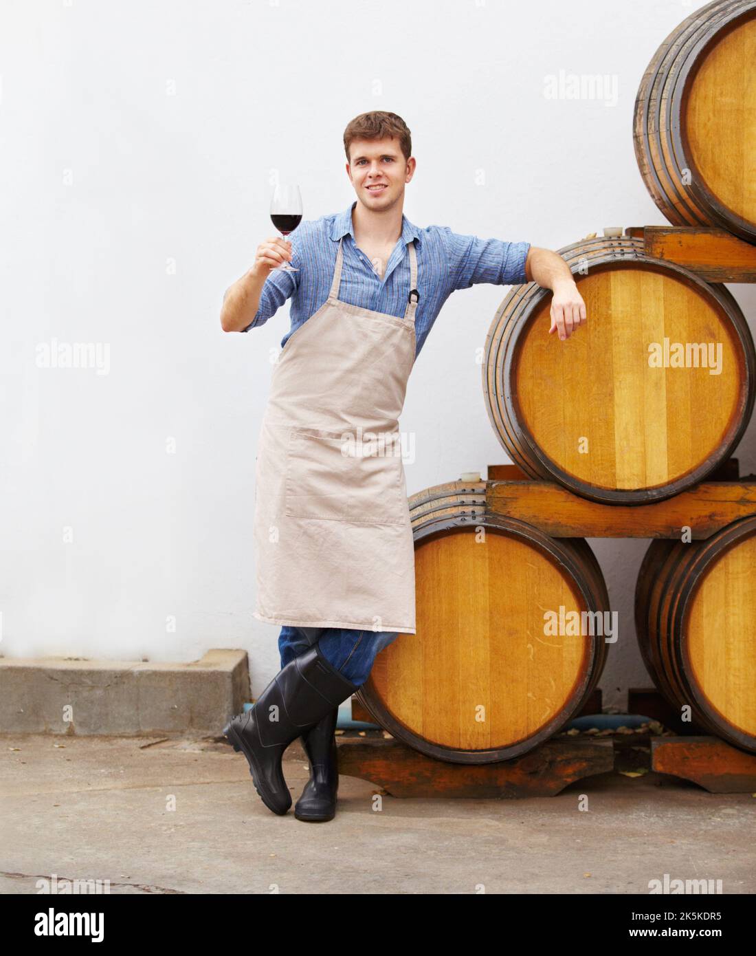 Genießen Sie einen feinen Rotwein. Ein junger Weinbauer hält ein Glas Rotwein in der Hand, während er sich auf ein Fass lehnt. Stockfoto