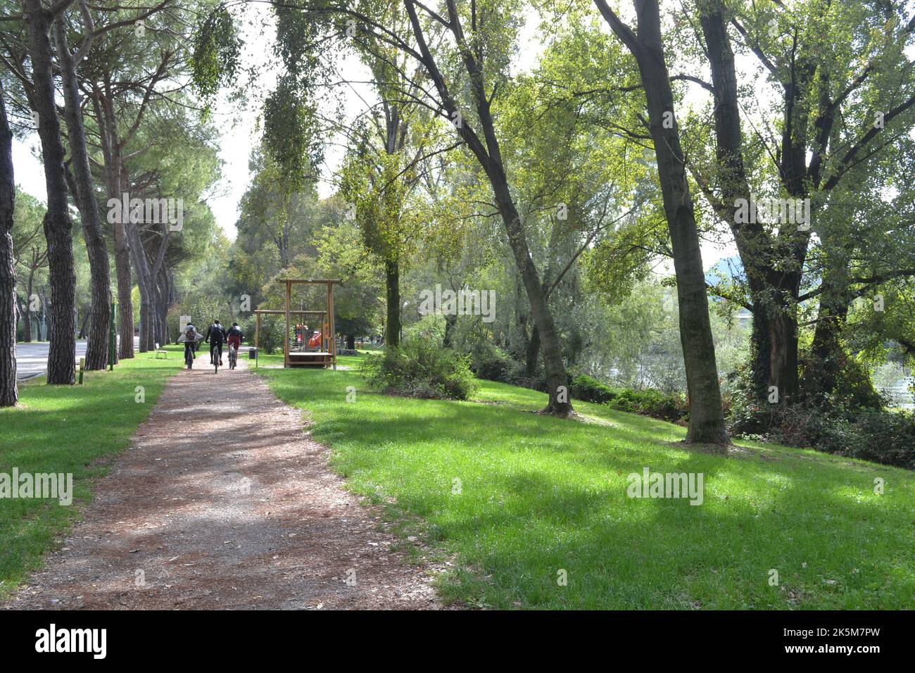 Lecco, Italien - 1. Oktober 2022: Landschaft mit breiten sandigen Gehweg im grünen Stadtpark mit Radfahrern in der Ferne. Radweg Lecco-Mailand. Stockfoto