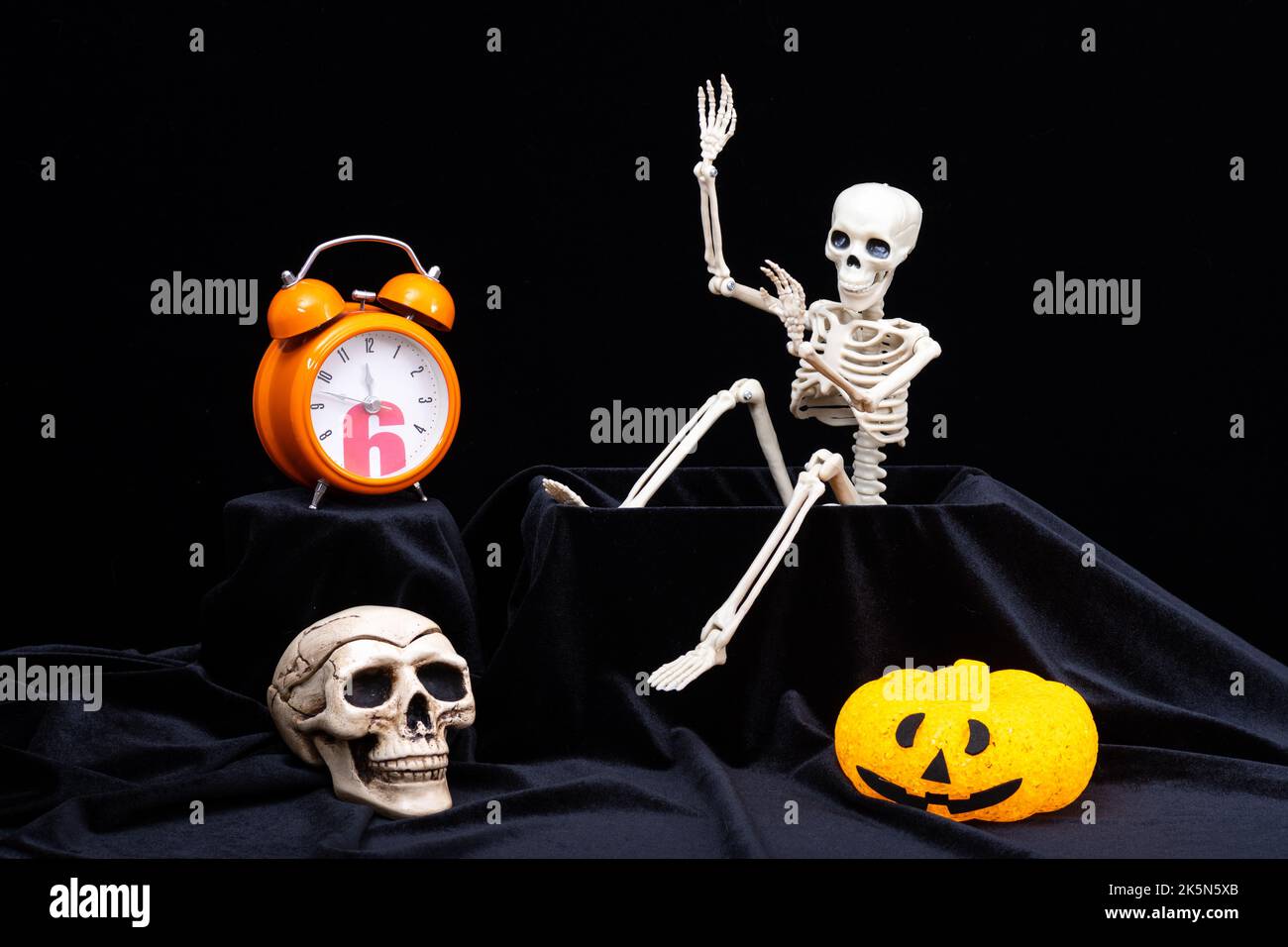 Halloween-Zeit. Ein Spielzeug-Skelett, das aus dem Grab kriecht und mit der Hand winkt, grüßt, Kürbisse mit Grimassen, eine orangefarbene Uhr, ein Totenkopf auf einem schwarzen Bac Stockfoto