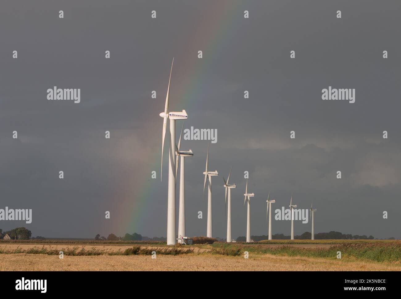 Eine Reihe von Windturbinen auf landwirtschaftlichem Gelände nahe der Küste, mit stürmischem Himmel und Regenbogen. Niederlande. 2022 Stockfoto