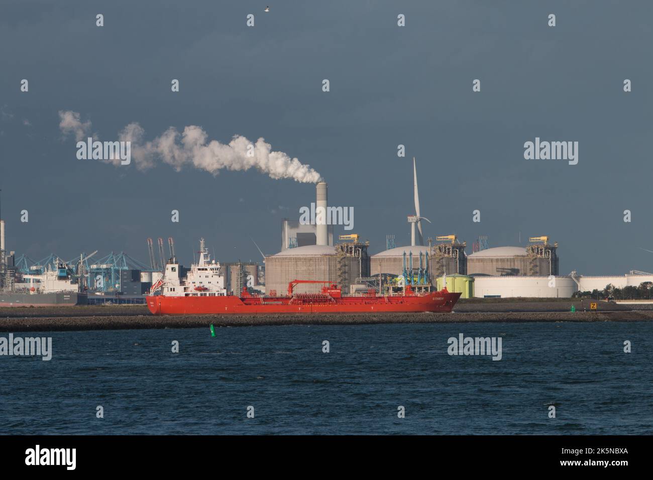 Schiff bewegt sich auf den Wasserstraßen mit Hafen im Hintergrund mit Schornstein emittierende Dampf oder Rauch. Vom Hook of Holland aus gesehen. Niederlande. 2022 Stockfoto