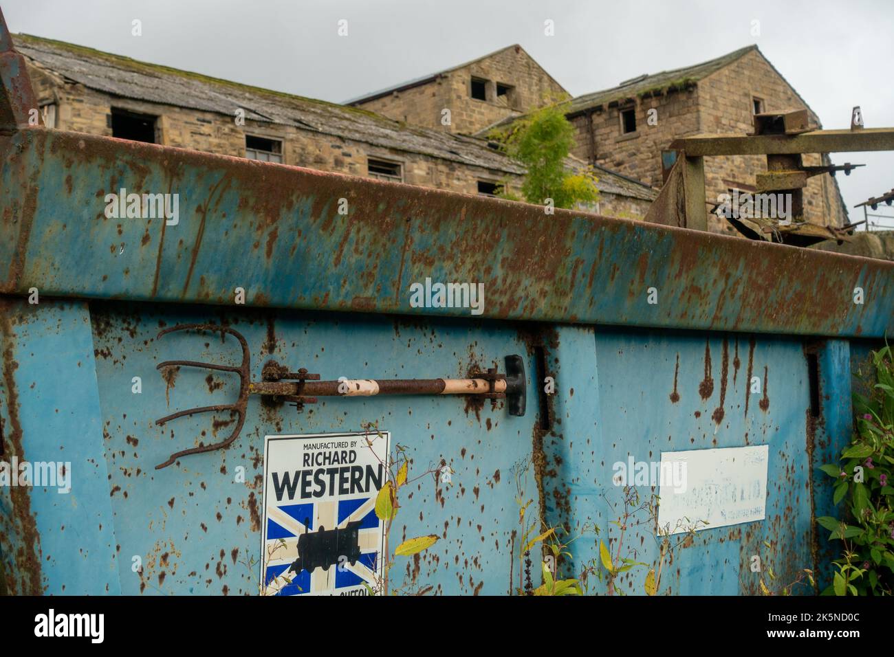 Farmer's Pitch Gabel an der Seite von rostigen Landmaschinen mit landwirtschaftlichen Gebäuden hinter, West Yorkshire, England, Großbritannien Stockfoto