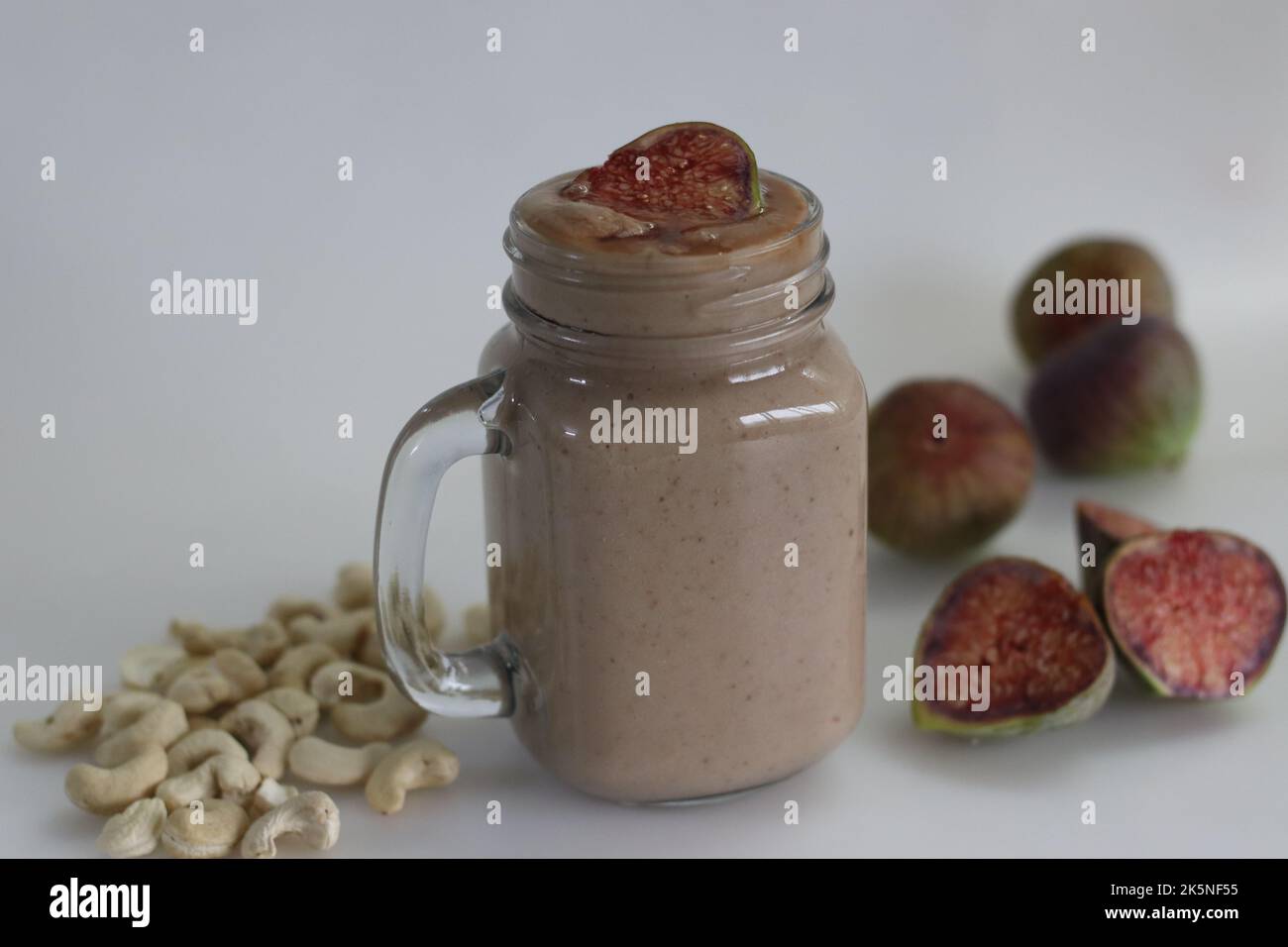 Fig Smoothie. Sommergetränk aus frischer Feige in Cashew-Milch, serviert in Glas-Smoothie-Glas. Aufgenommen auf weißem Hintergrund zusammen mit frischen reifen Feigen und cas Stockfoto