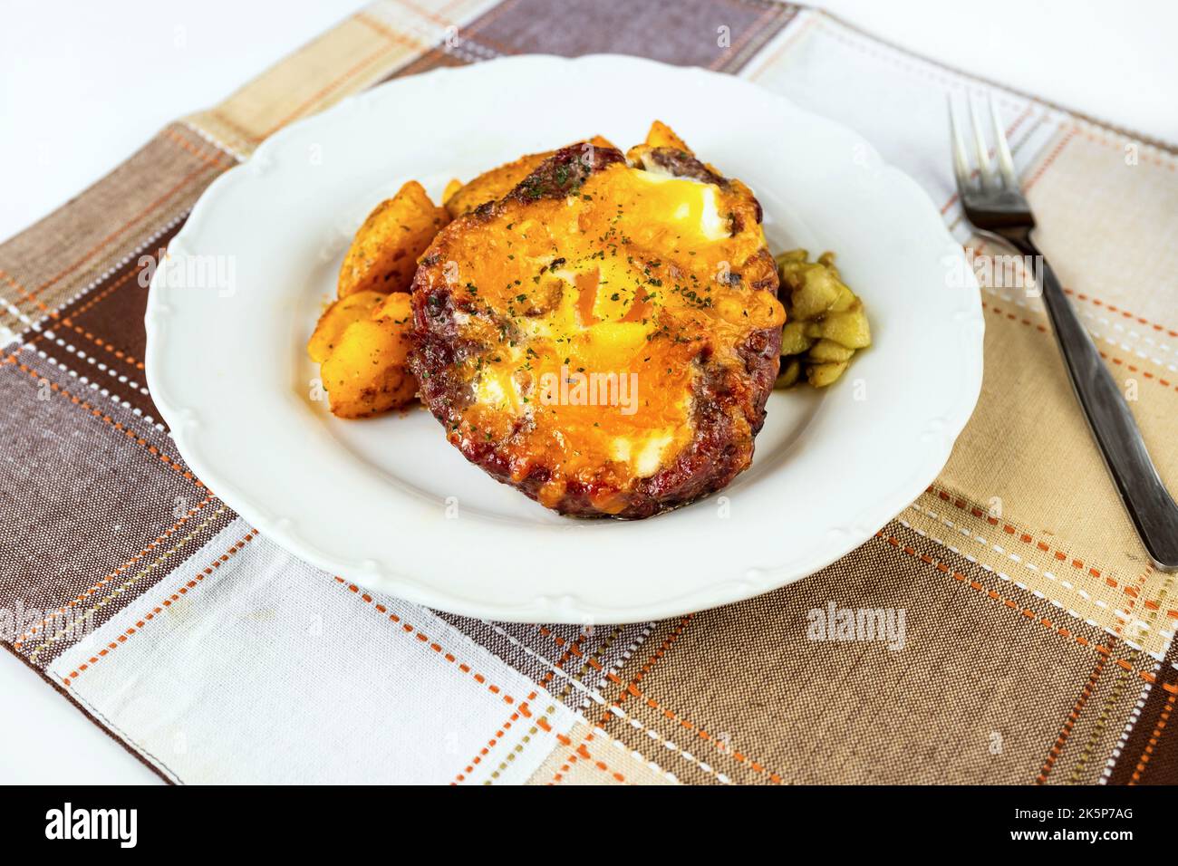 Rinderburger mit Spiegelei, Bratkartoffel und eingelegter Gurke auf weißem Teller auf kariertem Tischtuch auf dem Tisch. Stockfoto