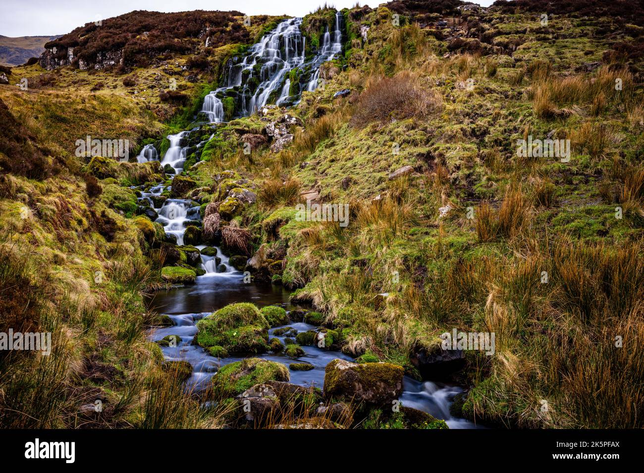 Ein wunderschöner Wasserfall, der die mit Moos bedeckten Felsen mit Bergen in der Ferne hinunterfließt Stockfoto