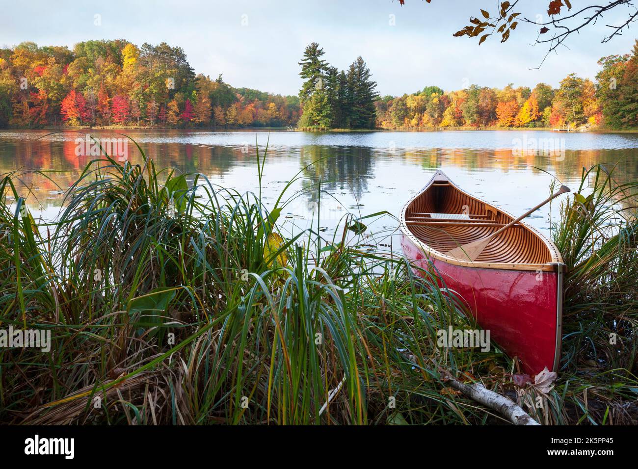 Rotes Holzkanu am Ufer des Sees mit Bäumen in Herbstfarbe und einer kleinen Insel Stockfoto