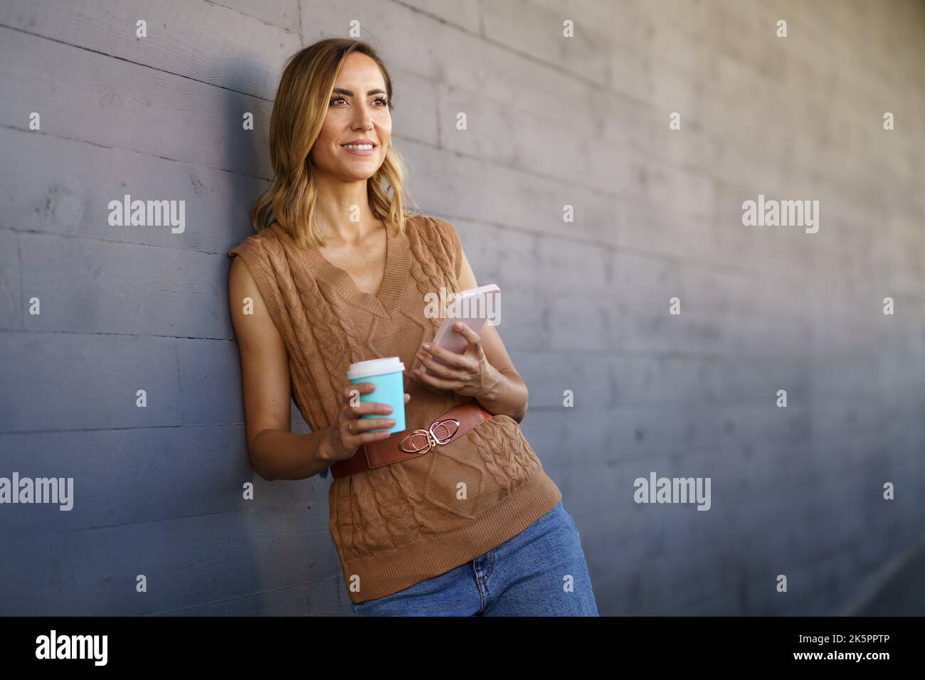 Frau mittleren Alters, die eine Kaffeepause nimmt. Kaukasische Frau, die einen Papierbecher benutzt und ihr Smartphone in der Hand trägt. Stockfoto