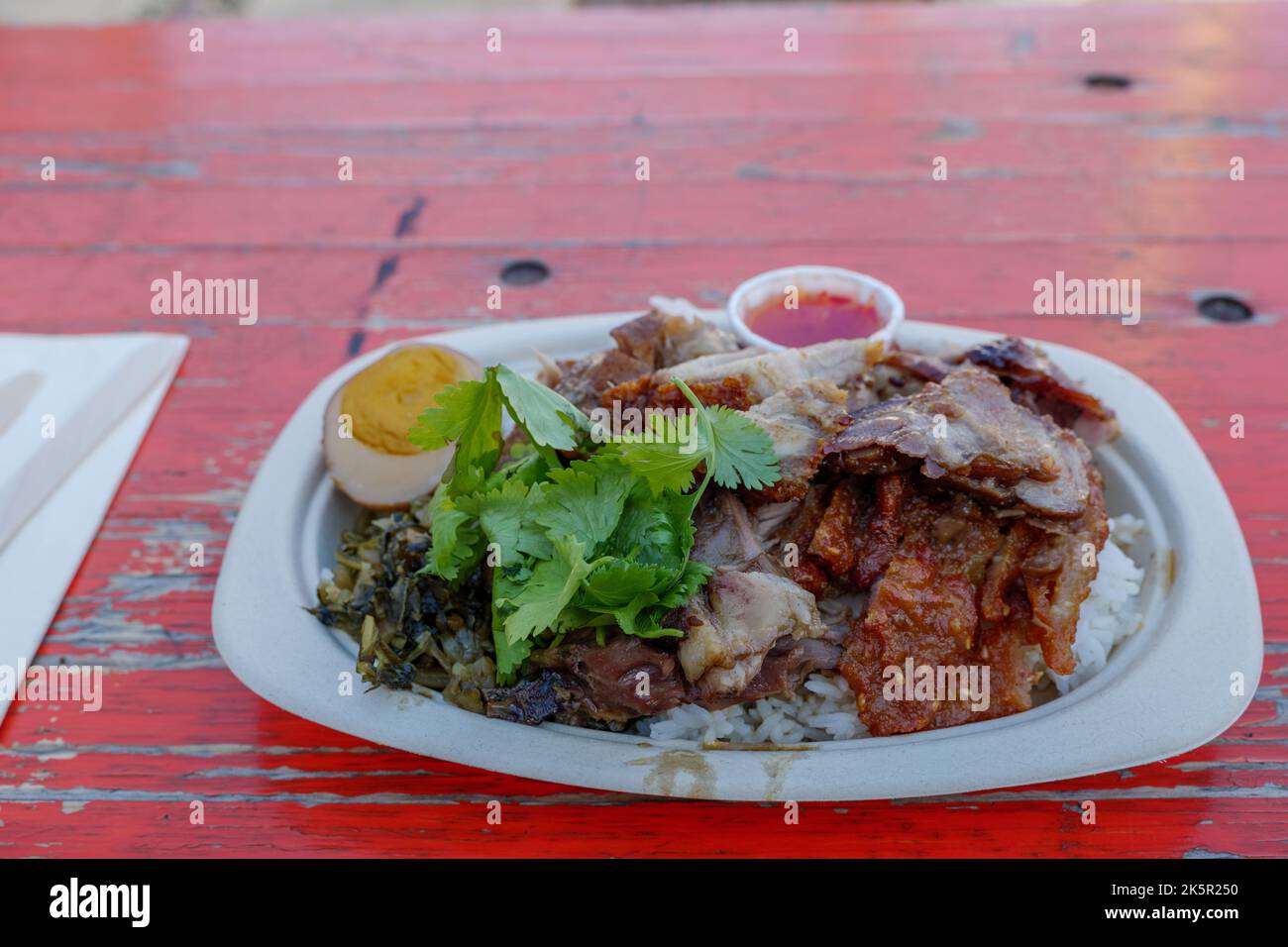 Geschmortes Schweineschenkel und Ei mit verschiedenen gepflückten Gemüse und gedämpftem Reis, auf einer Papierschale und einem rauen Holztisch. Stockfoto