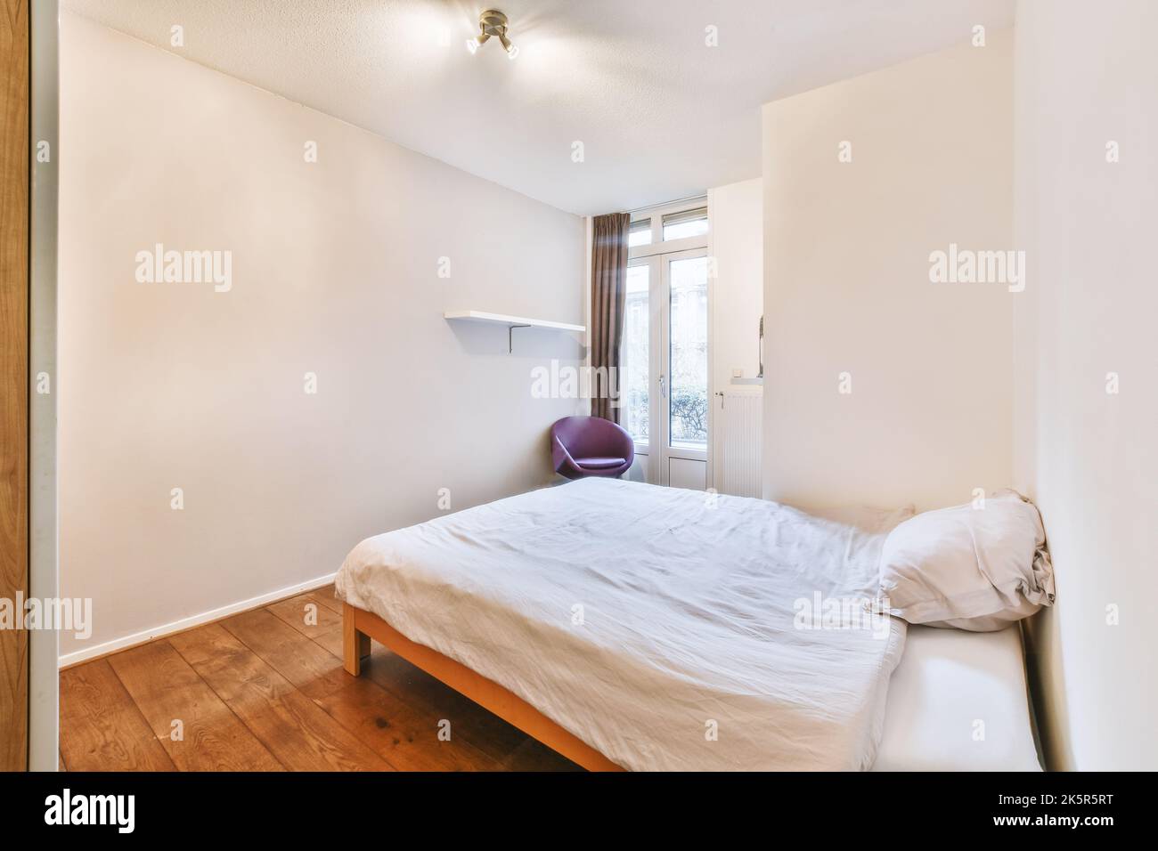 Komfortables Bett mit dekorativer Decke in der Nähe der Glastür und des Kleiderschrankes im hellen Schlafzimmer Stockfoto