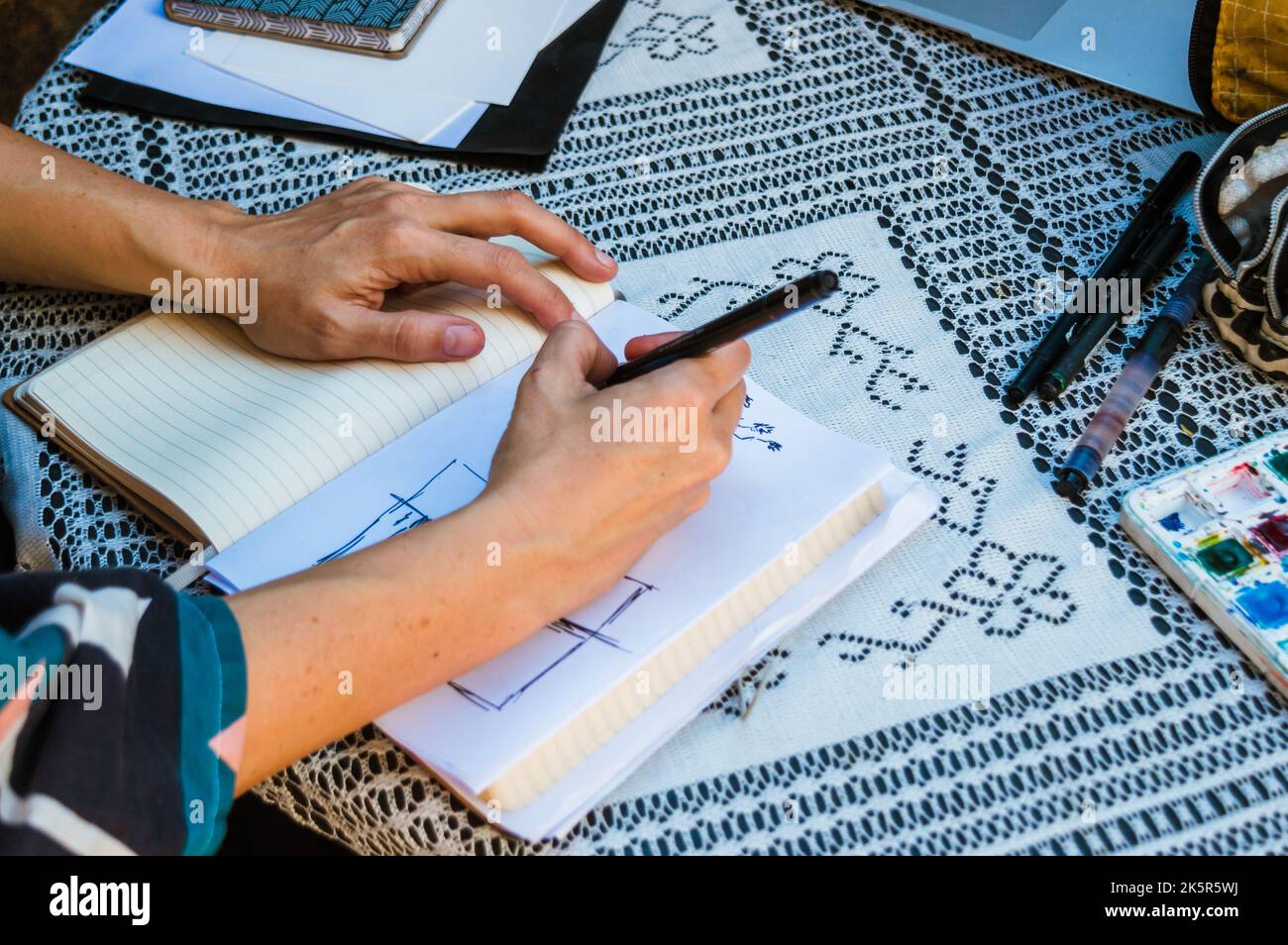 Weibliche kaukasische Hände im Freien auf einem Tisch zeichnen Skizzen auf einem weißen Blatt mit schwarzem Marker, mit Zeichengeräten auf dem Tisch, kopieren Raum. Stockfoto