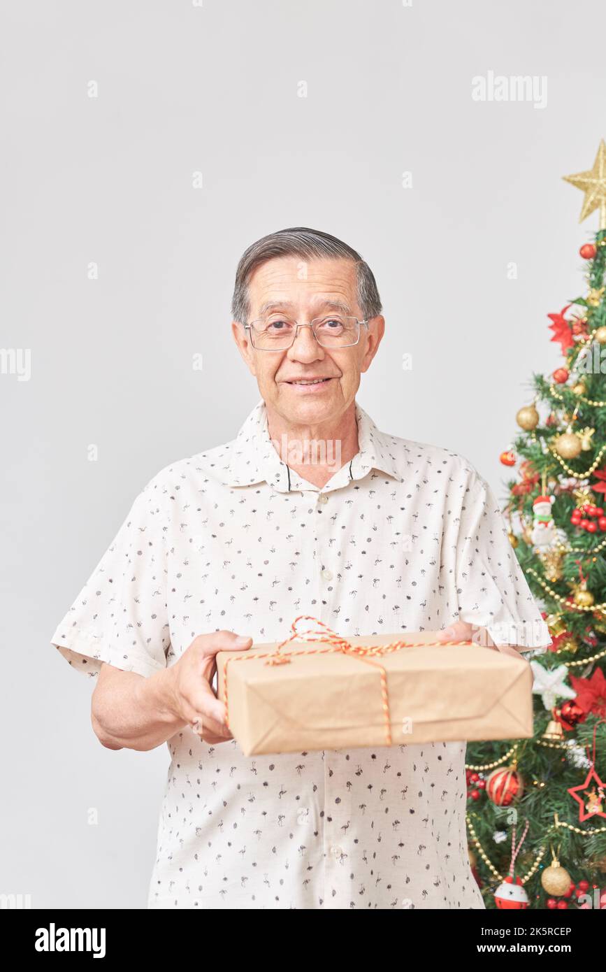 Ein älterer hispanischer Mann lächelt, als er der Kamera ein Weihnachtsgeschenk überreicht. Konzept: Die Freude am Schenken während der Feiertage. Vertikal komposit Stockfoto